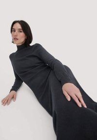 Dunkelgraues Strickkleid mit hohem Ausschnitt und langen Ärmeln. Das Material wirkt weich und hat eine fließende Silhouette sowie minimale Designdetails.