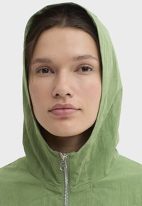 Veste à capuche verte avec une fermeture éclair frontale, fabriquée en tissu texturé, présentant un design simple et une capuche encadrant le visage.