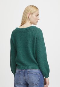Grüner Strickpullover mit lockerer Passform, weitem Rundhalsausschnitt, gerippten Bündchen und Saum, strukturiertem Stoff und langen Ärmeln. Getragen mit blauen Jeans.
