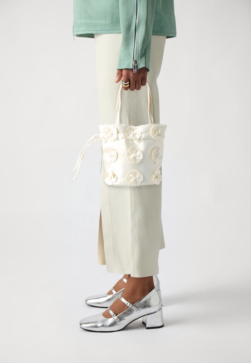 Femme tenant un sac seau blanc avec des ornements floraux, portant des chaussures à talons blocs argentés et une jupe et une veste de couleur claire.