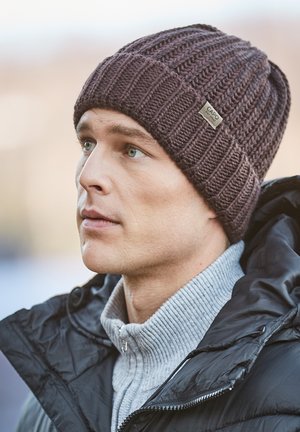 Mann trägt eine dunkelbraune gestrickte Mütze und eine schwarze Steppjacke über einem hellgrauen Rollkragenpullover, schaut im Freien zur Seite.