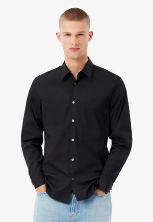 Jeune homme aux cheveux blonds courts portant une chemise noire à boutons et un jean bleu clair, les mains dans les poches, faisant face vers l'avant.