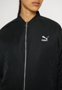 Personne portant une veste Puma noire avec une fermeture éclair sur le devant et un logo Puma blanc sur la poitrine gauche, cadrée au niveau du cou et des épaules.