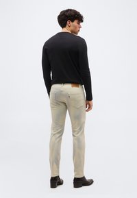 Νεαρός άνδρας που φοράει μπεζ τζιν στενής εφαρμογής, μαύρο μακρυμάνικο πουκάμισο και μαύρες μπότες, στέκεται με την πλάτη προς τα έξω σε απλό φόντο.