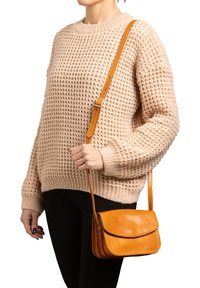 Piccola borsa a tracolla in pelle texturizzata di un caldo arancione, con chiusura a patta e tracolla regolabile, tenuta da una persona che indossa un maglione lavorato a maglia beige.