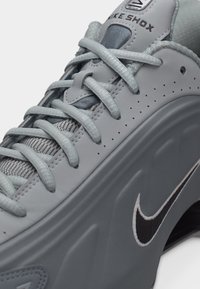 Prim-plan al unui pantof sport Nike Shox gri, prezentând șireturile, partea superioară perforată, limba căptușită și logo-ul negru Nike pe partea tălpii.