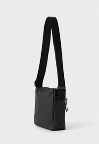 Μαύρη τσάντα ταχυδρόμου με κομψό, λείο φινίρισμα, ρυθμιζόμενο λουρί και κλείσιμο με φερμουάρ. Διαθέτει μινιμαλιστικό σχεδιασμό και γωνιακές άκρες.