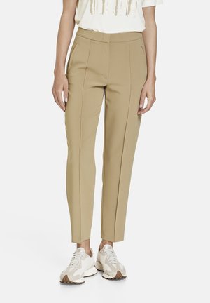 Personne portant un pantalon beige slim et des baskets blanches, debout devant un fond clair uni.