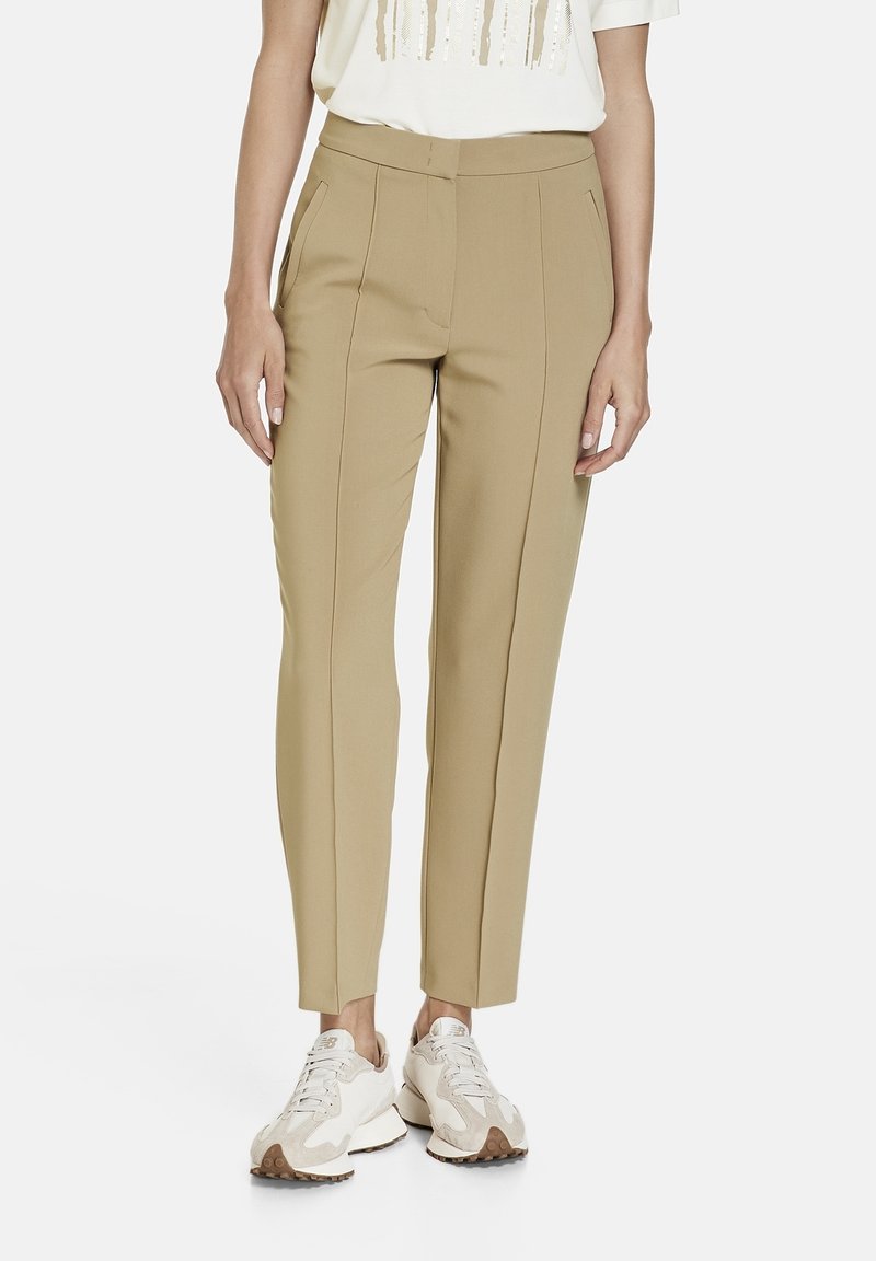Personne portant un pantalon beige slim et des baskets blanches, debout devant un fond clair uni.