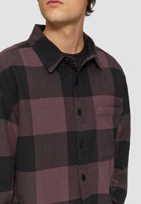 Chemise à carreaux prune et noir avec un design boutonné, un col et une poche poitrine. Fabriquée en tissu texturé, elle présente des boutons noirs.