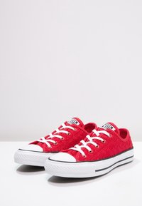 Zapatillas de lona rojas con textura acolchada, puntera de goma blanca y una franja negra a lo largo de la suela. Presentan cordones y ojales blancos.