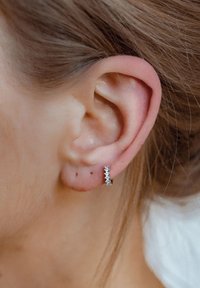 Aro plateado con cinco pequeñas piedras preciosas transparentes incrustadas en la parte frontal. La oreja está sin adornos, con piel suave y cabello castaño cercano.