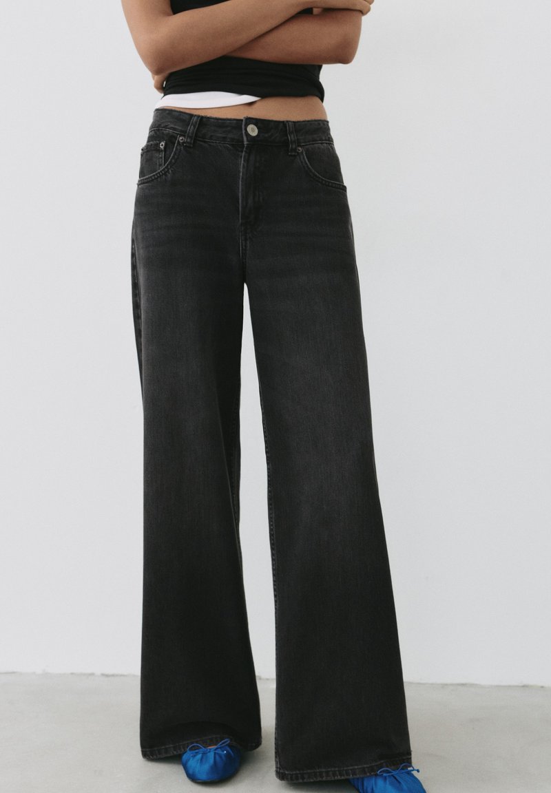 Personne portant un jean noir taille haute à jambes larges et des ballerines bleues, debout bras croisés devant un fond blanc uni.