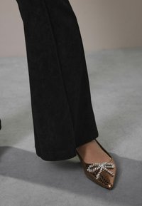 Woman wearing dark wide-leg pants and bronze pointed-toe flat shoes with rhinestone bow detail on gray floor.