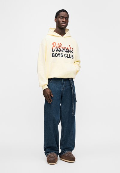 Κίτρινο φούτερ με κουκούλα με το κείμενο «Billionaire Boys Club», σε συνδυασμό με χαλαρά σκούρα τζιν και καφέ παπούτσια τύπου slip-on με κέντημα.