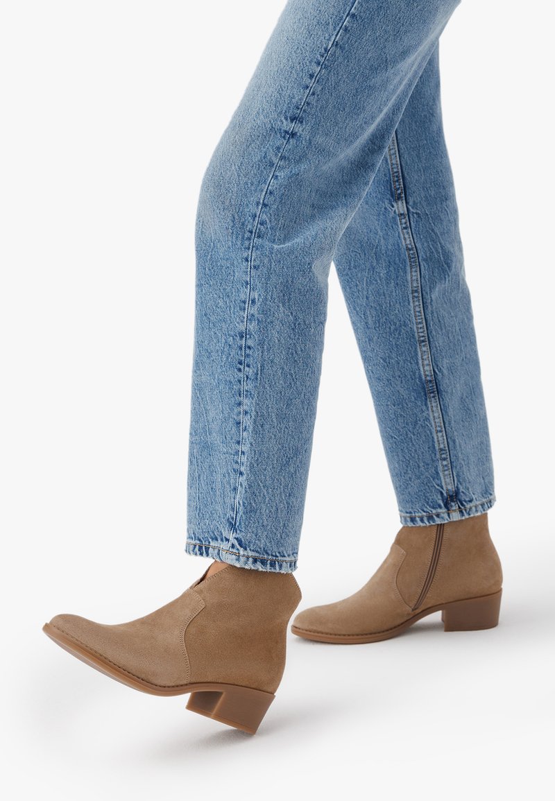 Jasnobrązowe zamszowe botki za kostkę na niskim słupku, noszone z jasnoniebieskimi dżinsami na białym tle.