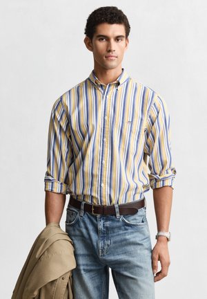 Giovane uomo con camicia a righe gialle, blu e bianche con maniche arrotolate, jeans azzurri, che tiene una giacca beige in una mano.