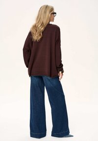 Brązowy oversize sweter z długimi rękawami, połączony z szerokimi jeansami w kolorze niebieskim. Gładka faktura i luźny krój widoczne z tyłu.