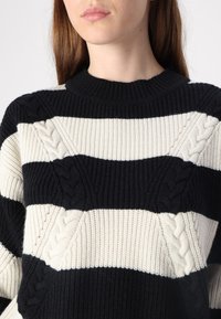 Jersey de punto con rayas horizontales en negro y crema, con patrones de cable texturizados en las secciones negras y un cuello de canalé.