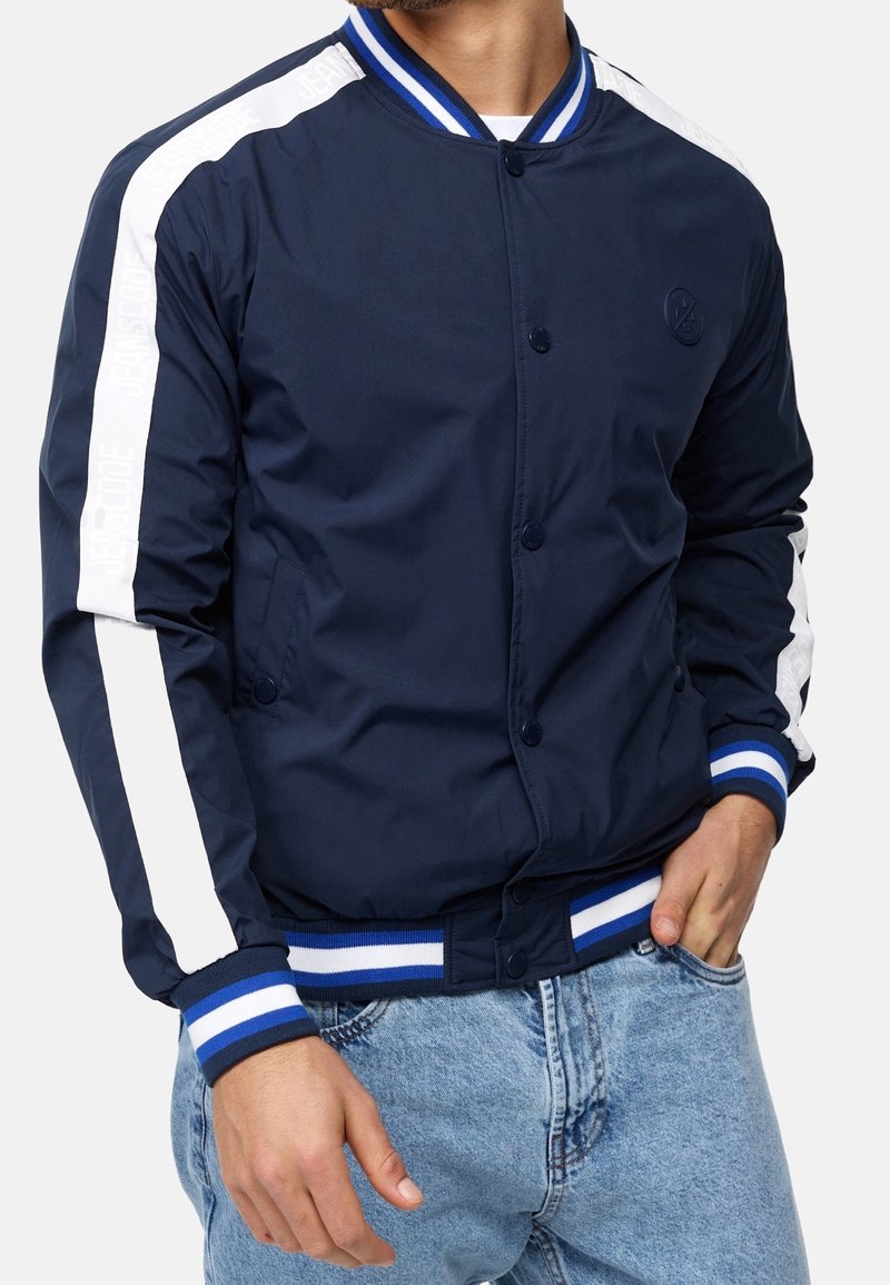 Homme portant une veste bomber bleu marine avec des poignets et un col rayés blancs et bleus, une bande blanche sur les manches, associée à un jean bleu clair.