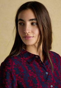 Młoda kobieta z długimi ciemnymi włosami, ubrana w koszulę w kwiatowy wzór w czerwono-niebieskie kolory, patrzy w lewo na tle beżowym.