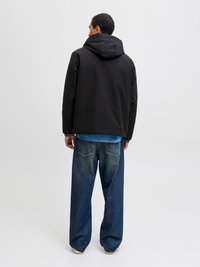 Μαύρο μπουφάν με κουκούλα και ματ φινίρισμα, συνδυασμένο με φαρδιά μπλε τζιν παντελόνια και ανοιχτόχρωμα υποδήματα, διακρίνεται από πίσω.