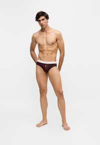Joven hombre atlético de pie descalzo, vistiendo calzoncillos rojo oscuro con cinturilla blanca sobre un fondo liso y claro.