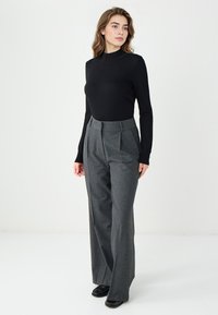 Zwarte lange mouwen top gecombineerd met hoog-geroemde, geplooide grijze broek met een gestructureerd patroon en zwarte schoenen. Effen achtergrond.