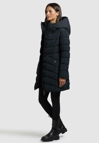 Schwarze Pufferjacke mit Kapuze, mit einem gesteppten Muster, Frontreißverschluss und seitlichen Taschen. Getragen über schwarzen Leggings und Stiefeletten.