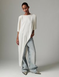 Λευκή, υπερμεγέθη τουνίκ με ασύμμετρο τελείωμα, συνδυασμένη με ανοιχτόχρωμο μπλε τζιν παντελόνι φαρδιάς γραμμής και μυτερά παπούτσια σε σχέδιο φιδιού.