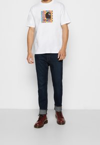 Hombre con camiseta blanca con el colorido logo de Carhartt, pantalones vaqueros oscuros arremangados y botas burdeos con cordones, de pie sobre un suelo gris.