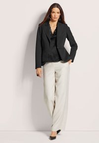 Schwarze Blazer mit tailliertem Design, Reverskragen und zwei Vordertaschen. Kombiniert mit einem schwarzen Satin-Top und hellgrauen weit geschnittenen Hosen.