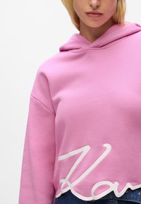 Rosa Sweatshirt mit Kapuze und weißem, wellenförmigem Schriftzug am Saum. Das Material sieht weich und strukturiert aus.