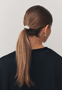 Schwarzes Haargummi mit einem silbernen, mit Strasssteinen verzierten Clip, das glattes braunes Haar zu einem Pferdeschwanz zusammenhält. Der Hintergrund ist neutral und schlicht.