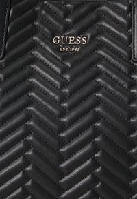 Sac à main structuré noir avec un motif en chevrons en matériau texturé. L'avant présente le logo "GUESS" en accents métalliques argentés.