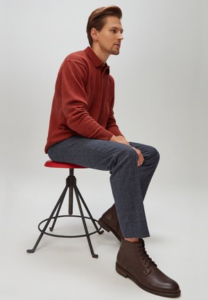 Brązowe skórzane sznurowane botki do kostki, granatowe spodnie w kratę oraz sweter polo w kolorze rdzy. Model siedzi na czerwonym stołku.