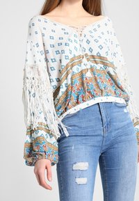 Biała bluzka w stylu boho z niebiesko-złotymi wzorami, obszernymi rękawami z frędzlami i dekoltem na sznurowadła, zestawiona z podartymi dżinsami.