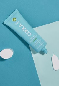 Hellblauer Tubus von COOLA Classic Face Sunscreen, SPF 30, mit grünen Akzenten. Weiße Sonnencreme wird herausgedrückt und auf einem pastellfarbenen Hintergrund platziert.