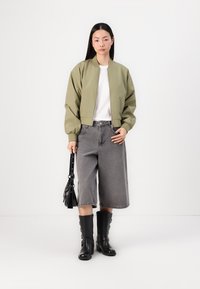 Olive green bomber jacket with ribbed cuffs, white t-shirt, grey wide-leg denim shorts, black knee-high boots, and a small black handbag.