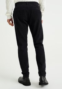 Černé kordové kalhoty s úzkým střihem, bočními kapsami a strukturovaným povrchem. Nosené s černými kotníkovými botami, na kterých jsou vidět elastické manžety.