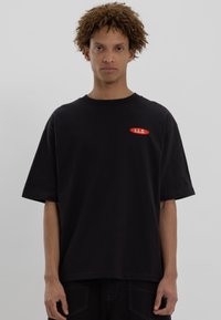 Black cotton t-shirt with a relaxed fit, short sleeves, and a red oval patch featuring the text "L.L.S." on the chest.