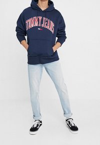 Felpa navale con logo "TOMMY JEANS" in rosso, tasca a marsupio; abbinata a jeans azzurri con risvolto e sneaker nere.