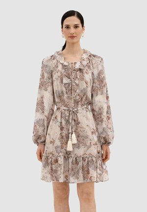 Donna che indossa un mini abito a maniche lunghe con fantasia floreale, volant, cintura con nappine e collo alto, in piedi contro uno sfondo neutro.