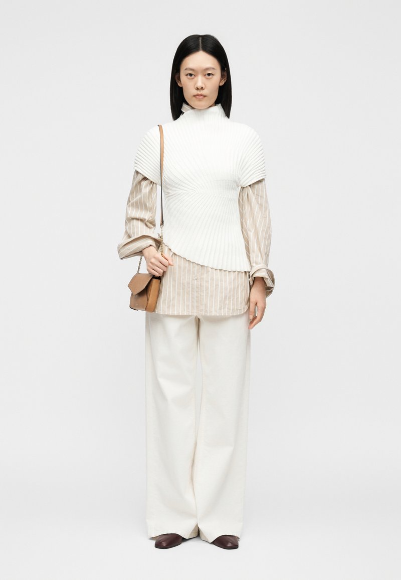 Γυναίκα με ίσια μαύρα μαλλιά που φοράει λευκό πλεκτό γιλέκο πάνω από μία μπεζ ριγέ πουκάμισο, λευκό παντελόνι με φαρδιά μπατζάκια, καφέ παπούτσια και κρατά μια ταμπά τσάντα.