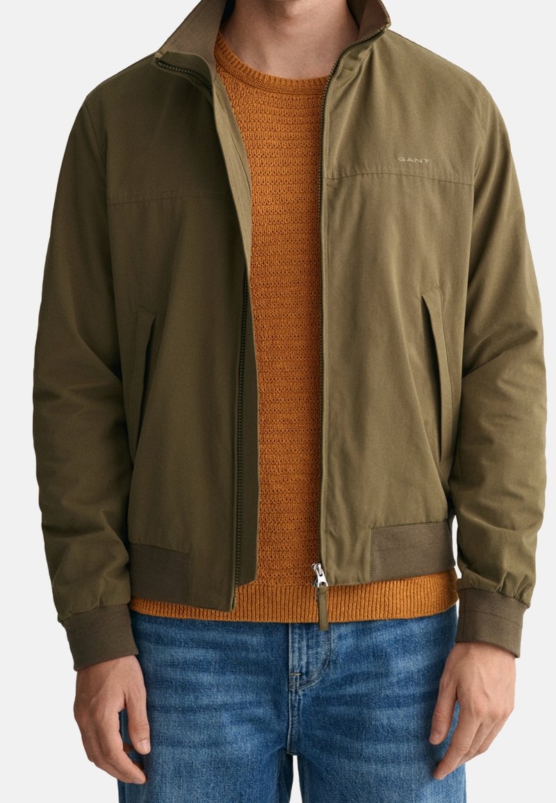 Homme portant une veste zippée vert olive par-dessus un pull en maille orange et un jean bleu, debout, les bras détendus le long du corps.