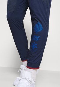 Navy Jogginghose mit strukturiertem Obermaterial, versehen mit blauen Druckgrafiken: einem Globus, "STAY LOST" und Richtungssymbolen. Rote Akzente an den Bündchen.
