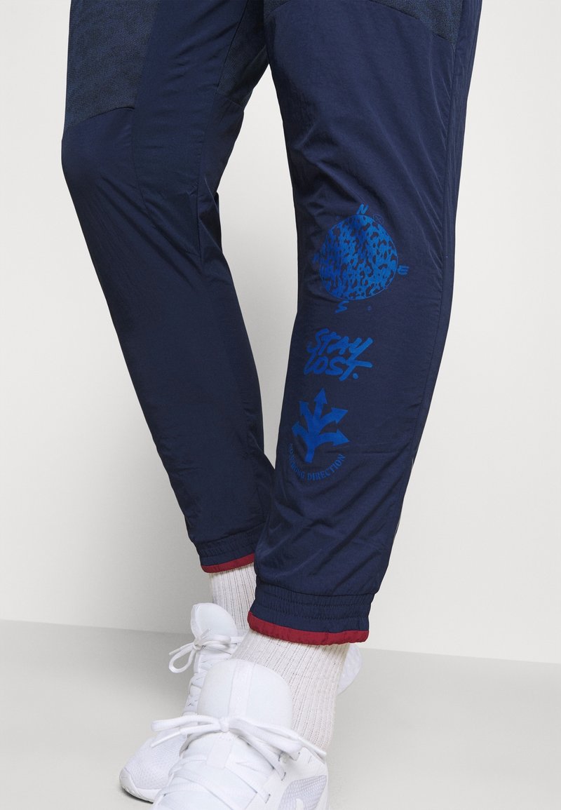 Navy Jogginghose mit strukturiertem Obermaterial, versehen mit blauen Druckgrafiken: einem Globus, "STAY LOST" und Richtungssymbolen. Rote Akzente an den Bündchen.