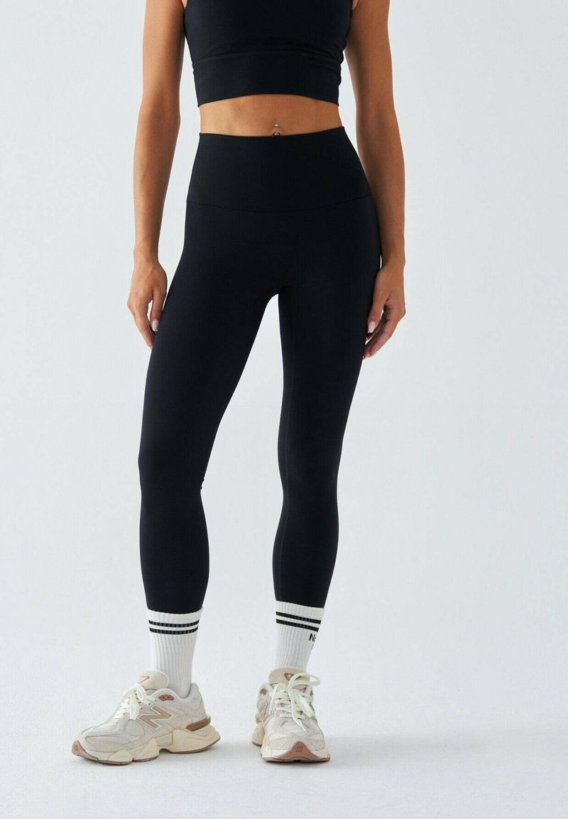 Czarne legginsy z wysokim stanem o gładkiej fakturze. Połączone z białymi skarpetkami z czarnymi paskami oraz beżowymi butami sportowymi z akcentami.