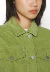 Chaqueta de pana verde con cuello, bolsillos frontales y botones plateados. La tela texturada presenta un ribeteado vertical.