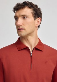 Rostfärgad polo skjorta med dragkedja vid kragen, tillverkad av ett mjukt tyg. Har en broderad logotyp på bröstet.